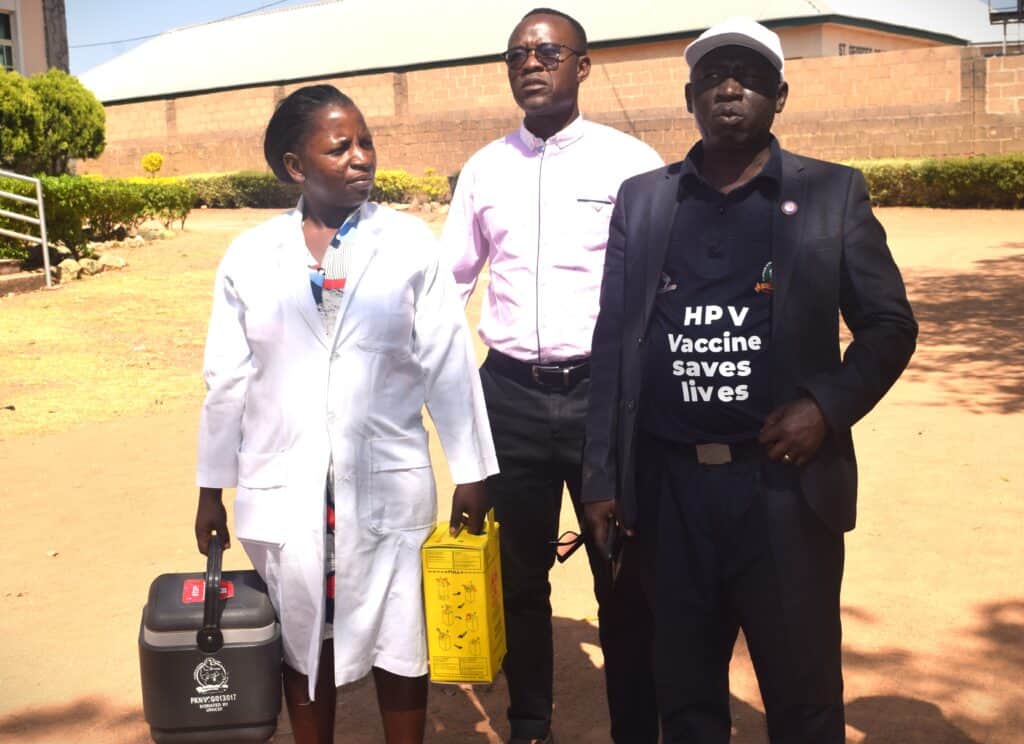 A nurse stands with two other people, holding supplies.