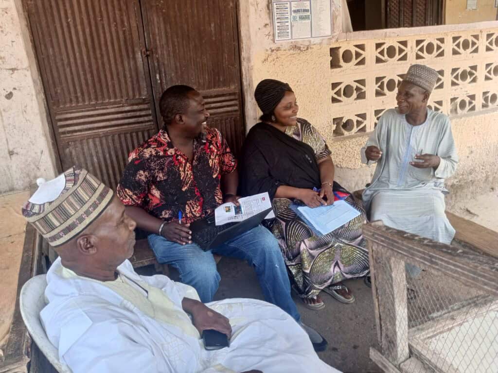 A Nigerian man sits with three people talking to them. The people are holding documents.