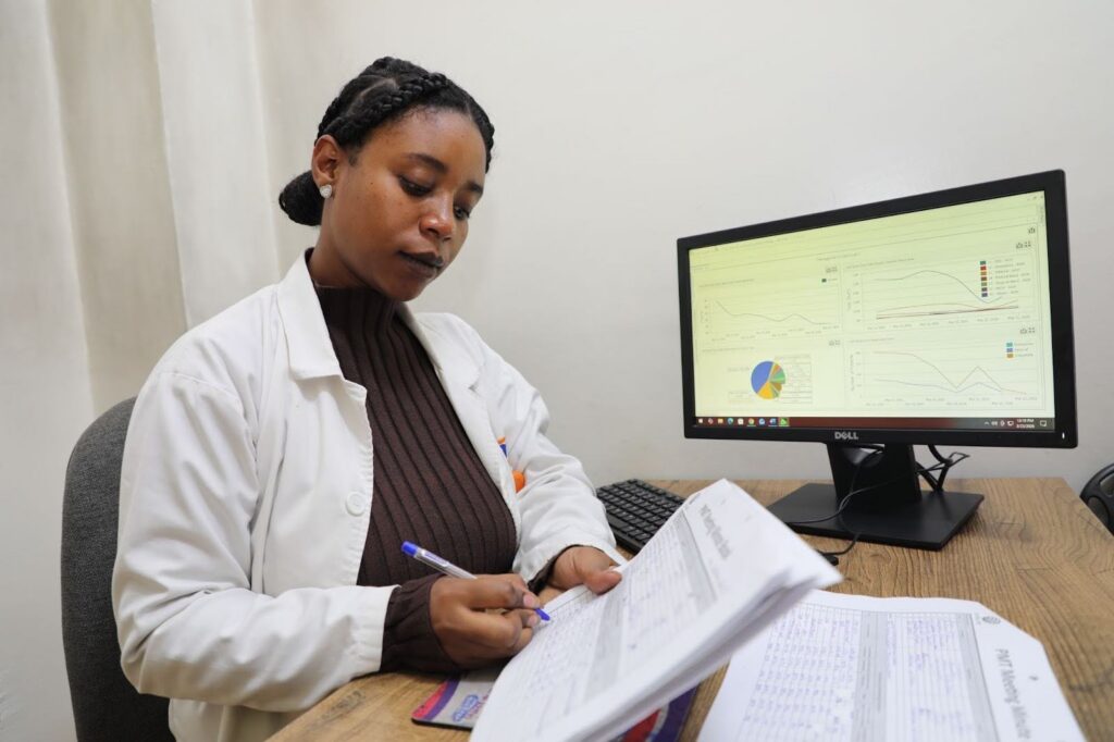 A woman in a white coat sits at a desk taking notes. The computer behind her shows graphs and charts on the screen.
