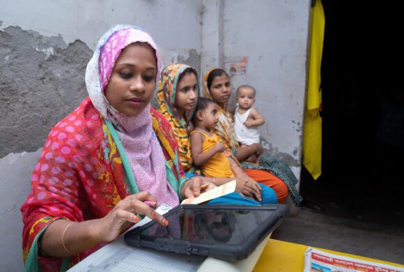 A female health care worker is inputting information into a tablet two women sit behind her holding children.