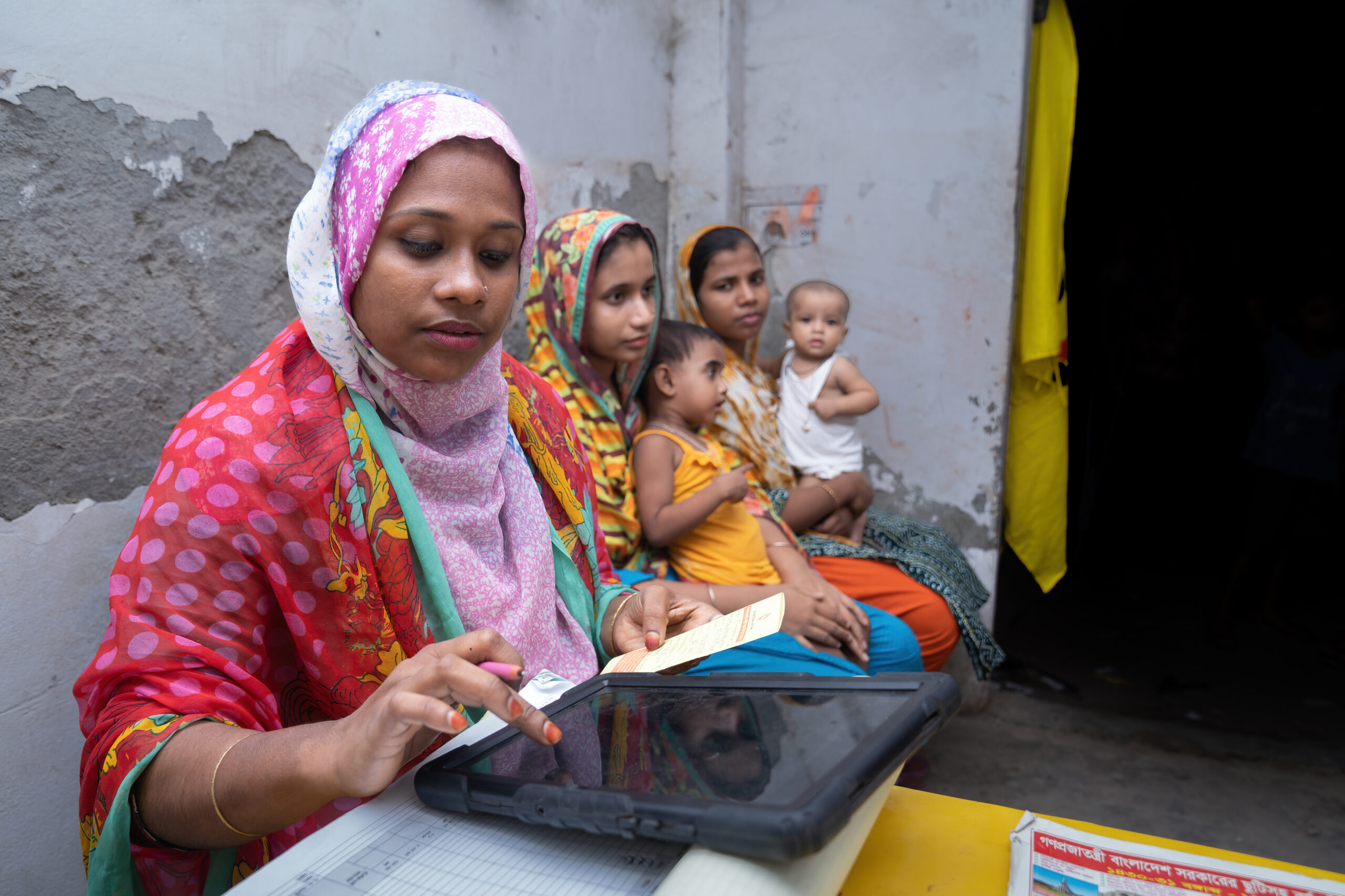A female health care worker is inputting information into a tablet two women sit behind her holding children.